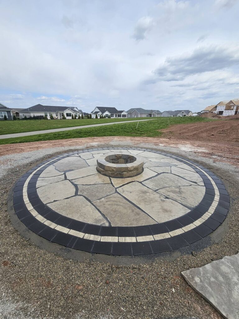 Circular flagstone patio with central stone fire pit and dark brick paver border in a backyard.