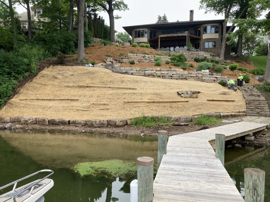 A multi-level tiered landscape design on a sloped waterfront property featuring natural stone retaining walls, lush green plantings, and a wooden dock leading to a modern stone home.