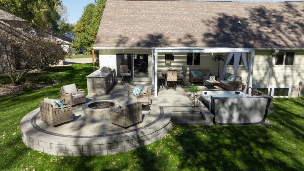 A multi-level backyard paver patio in Neenah, WI, featuring a raised circular fire pit seating area with stone retaining walls, an outdoor kitchen, and a white pergola-covered dining space.